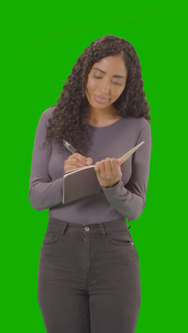 Vertical-Video-Portrait-Of-Female-College-Or-University-Student-Writing-In-Notebook-Against-Green-Screen-Smiling-At-Camera